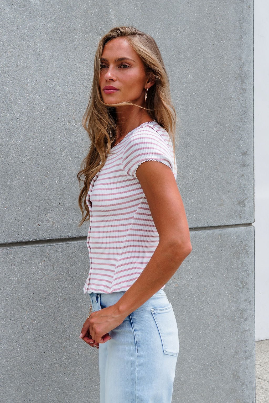 Woman with long hair wears the Free People Stripe Most Wanted Top - Mauve and light blue jeans, standing by a gray concrete wall.