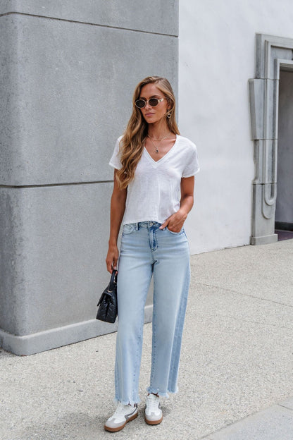 Woman in sunglasses, Free People Take 2 Tee - Ivory, light blue jeans, white shoes, stands outside holding a small black purse.