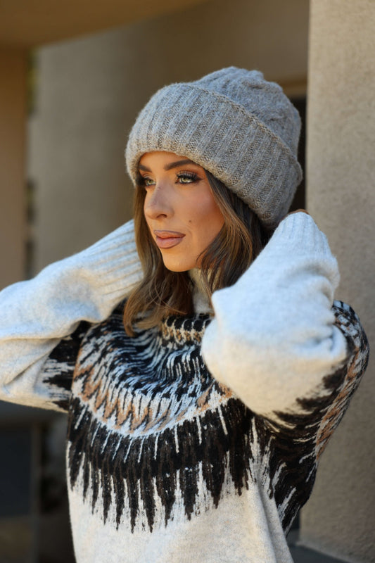 A woman outdoors adjusts her Free People Taupe Coastline Beanie, gazing thoughtfully to the side in a patterned sweater.