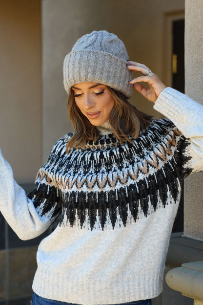 Woman in a knit sweater and Free People Taupe Coastline Beanie, looking down and touching her hat outdoors by a building.