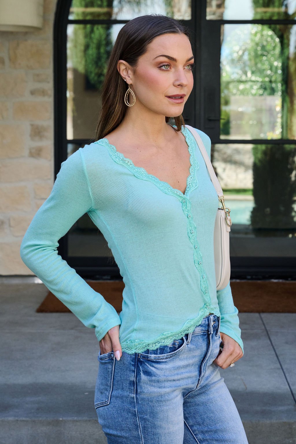 A woman in a Free People Teal Big Love Ribbed Top and jeans stands outdoors, holding a white purse and looking to the side.