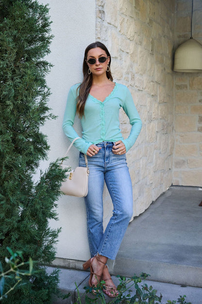 Woman in sunglasses poses by a wall in jeans, sandals, a Free People Teal Big Love Ribbed Top, and a white purse.