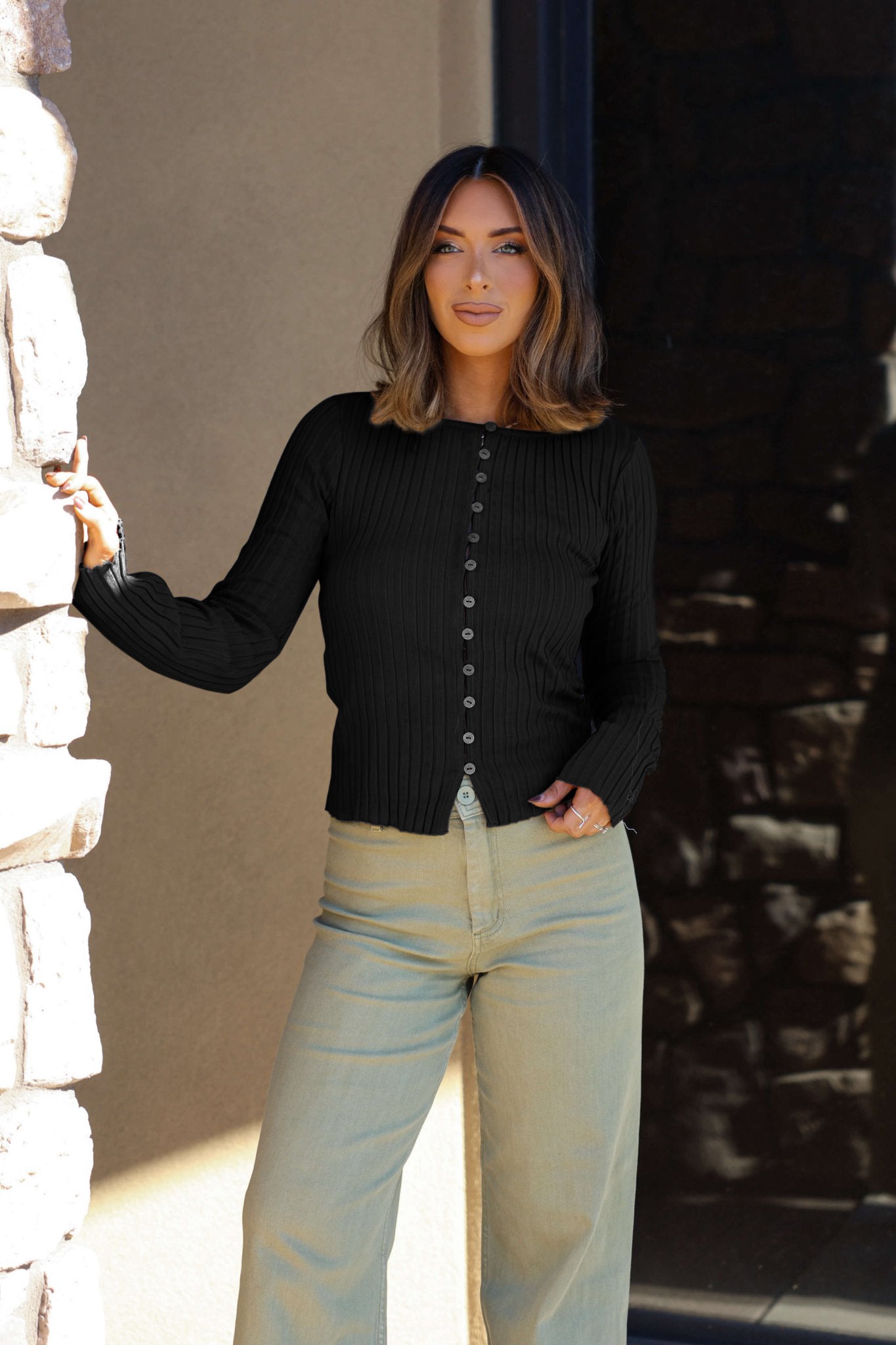 Woman with shoulder-length hair, smiling by a stone wall in a Free People Tilly Black Long Sleeve Top and light pants.