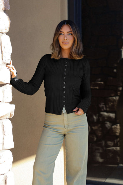 Woman with shoulder-length hair, smiling by a stone wall in a Free People Tilly Black Long Sleeve Top and light pants.