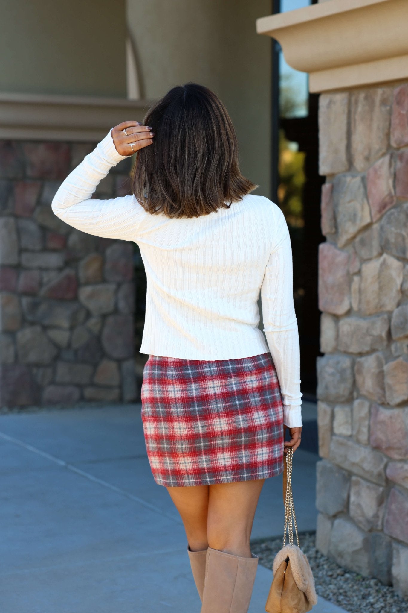 A woman with shoulder-length hair stands outside, wearing the Free People Tilly Ivory Long Sleeve Top and a plaid skirt, holding a beige purse.