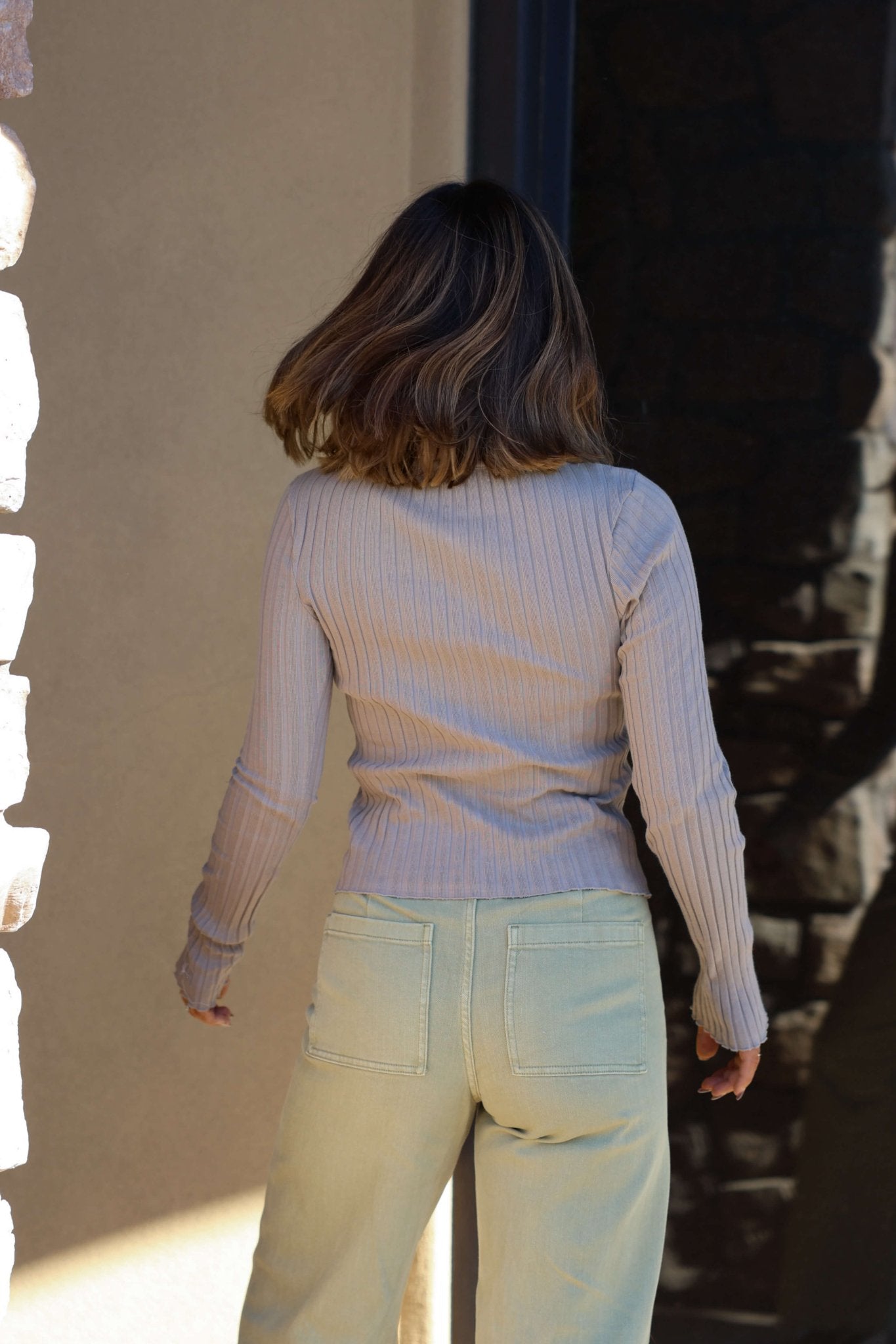 A woman in the Free People Tilly Taupe Long Sleeve Top stands with her back to the camera, wearing light jeans.