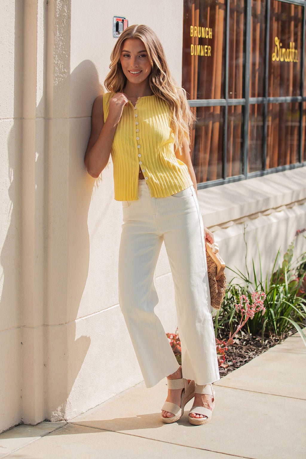 A woman in the Free People Tilly Yellow Vest and white pants smiles against a wall with plants and a restaurant window behind her.