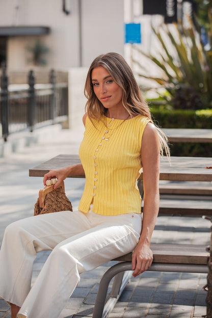 Wearing a Free People Tilly Yellow Vest and white pants, she sits outdoors holding a small woven bag—a chic wardrobe staple.