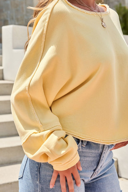 Wearing the Free People To The East Pullover in butter yellow and light blue jeans, a person stands on steps enjoying the sunlight.