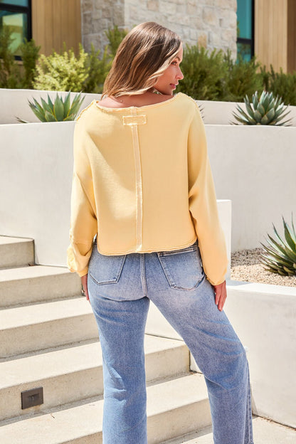 A woman stands on steps outdoors, facing away, in blue jeans and a Free People To The East Pullover in butter yellow.