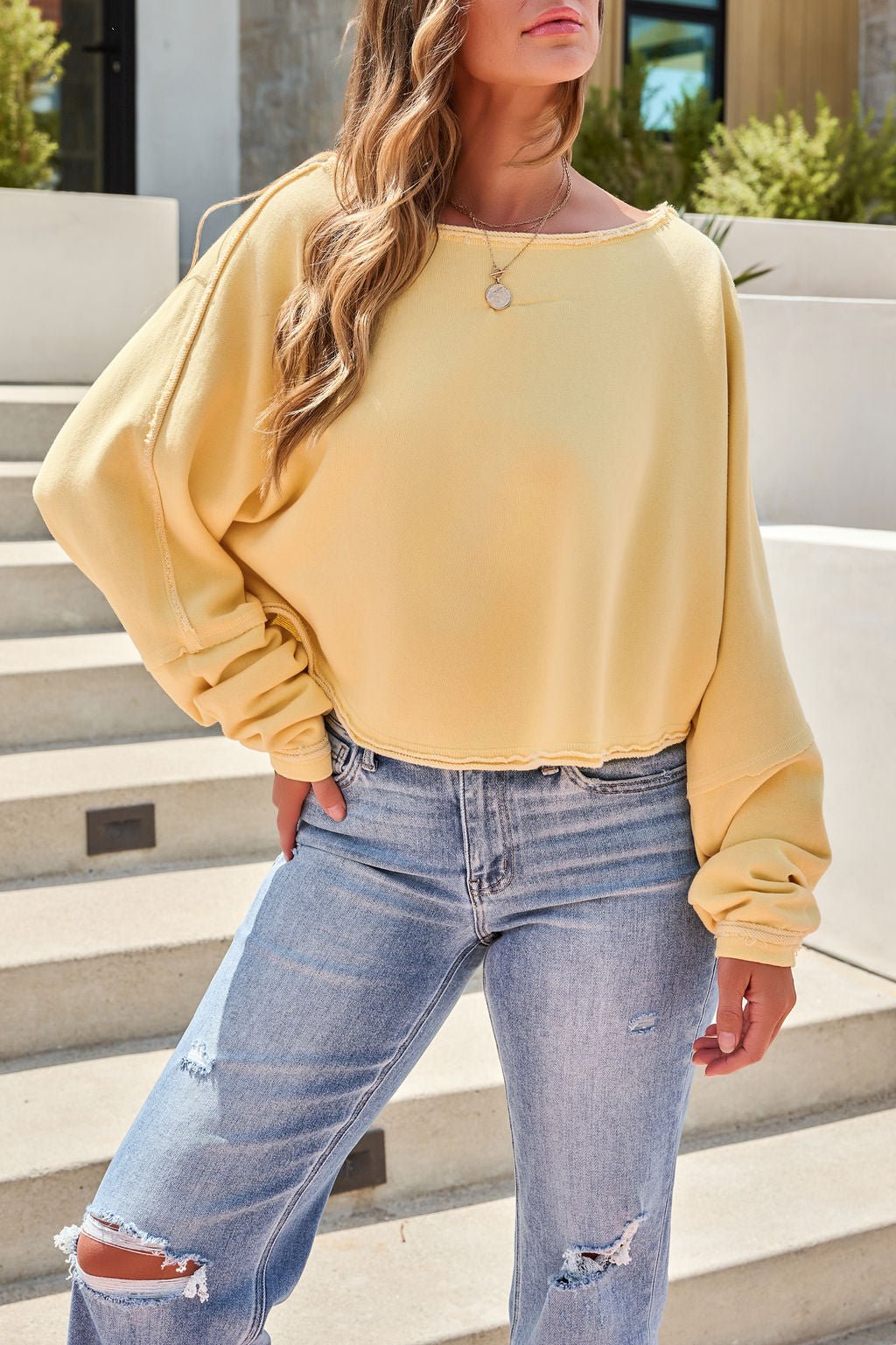 A woman in the Free People To The East Pullover in butter yellow and ripped jeans stands outside on concrete steps.
