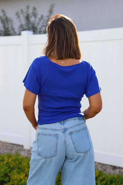 A woman in a Free People Violet Blue Lucy Off The Shoulder Tee and light blue jeans stands outdoors, facing away from the camera.