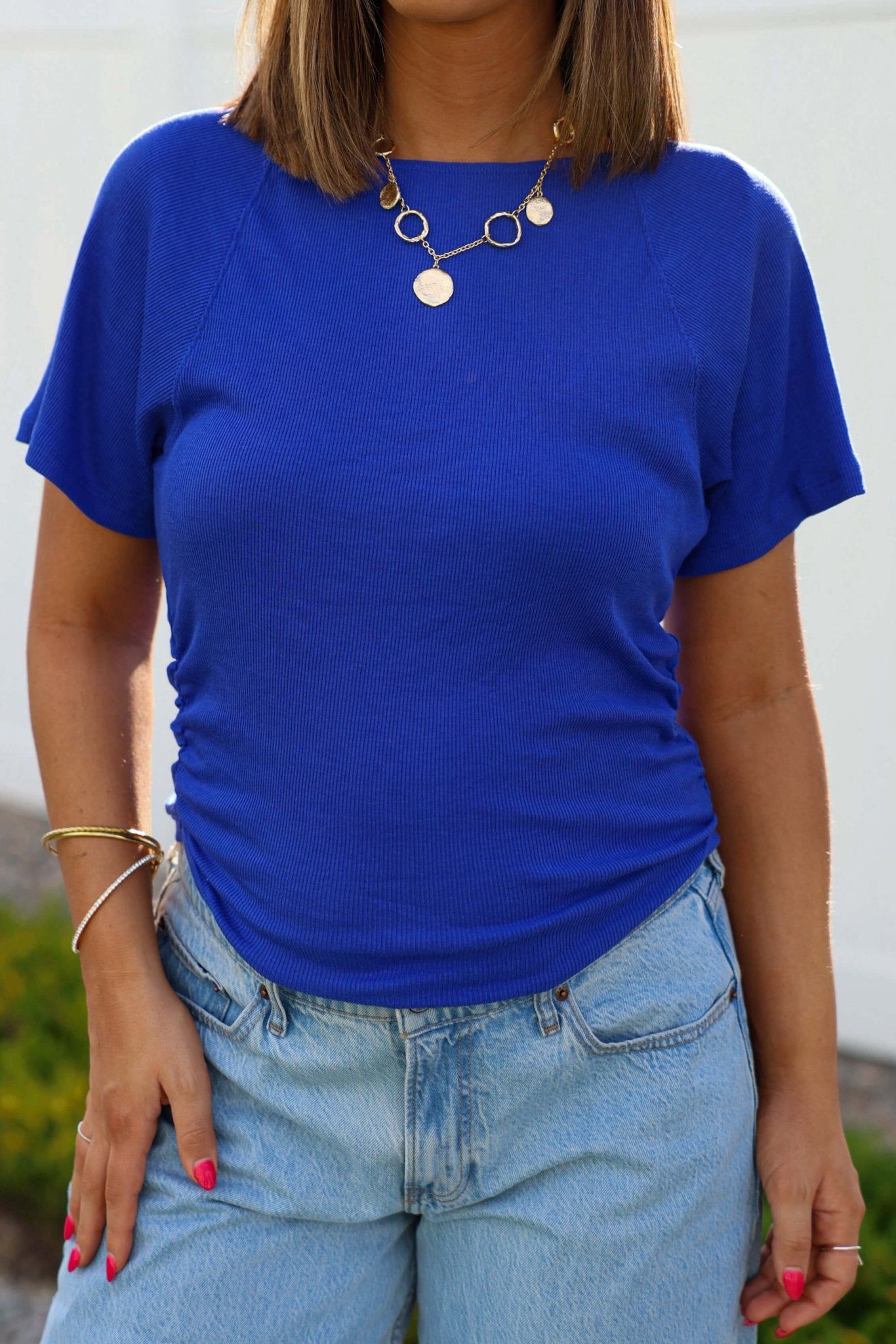 Woman outdoors in Free People Violet Blue Lucy Off The Shoulder Tee, light blue jeans, gold jewelry, and pink nails.