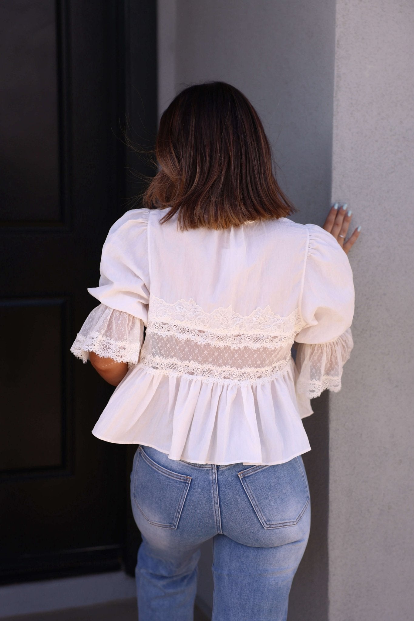 A woman in a white Free People Forever Young Top - FINAL SALE and light blue jeans stands facing a wall, her brown hair shoulder-length.