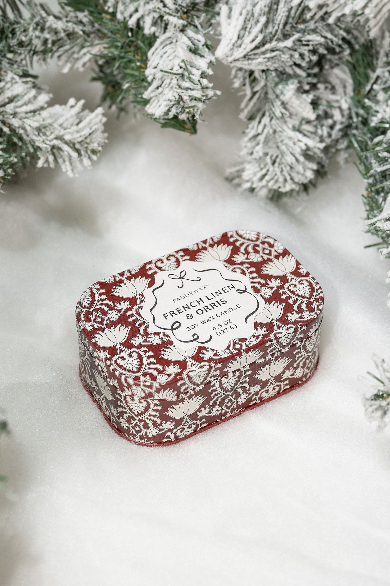 A red and white patterned soap bar sits on snow beside a French Linen & Orris Print Tin Soy Candle and snowy pine branches.
