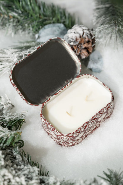 The French Linen & Orris Print Tin Soy Candle comes in a decorative tin with frosted pine, snow, and pinecones.