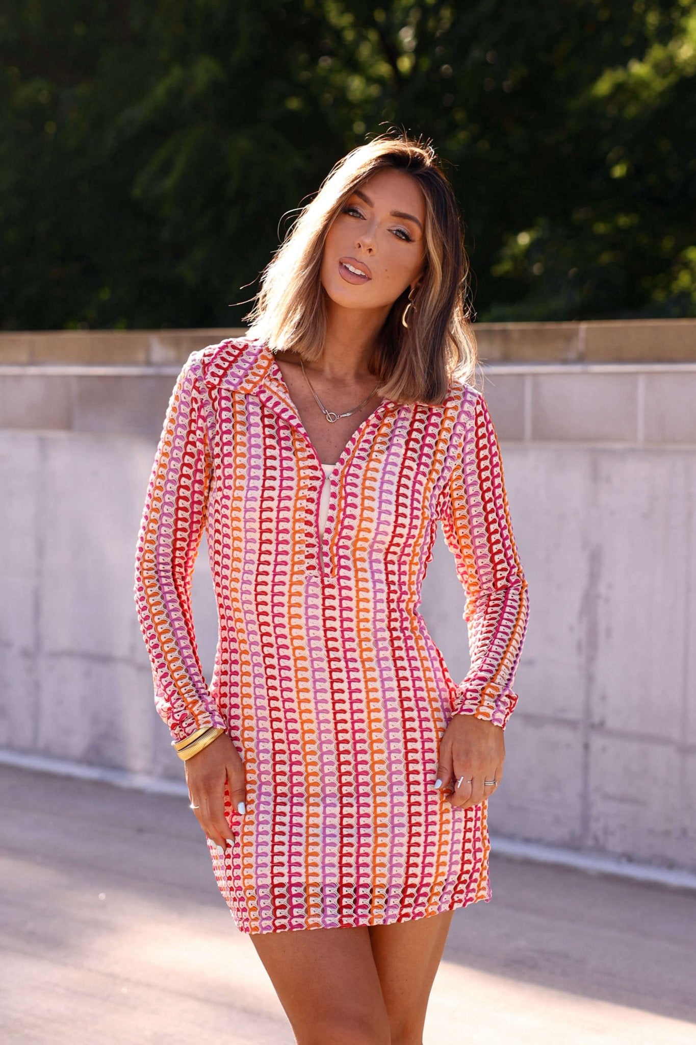 Woman in a Fuchsia Multi Striped Crochet Mini Dress stands outdoors by a concrete wall with greenery in the background.