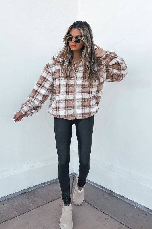Woman in sunglasses wears the Fuzzy Brown Plaid Zip Up Jacket with black leggings and beige shoes against a white wall.