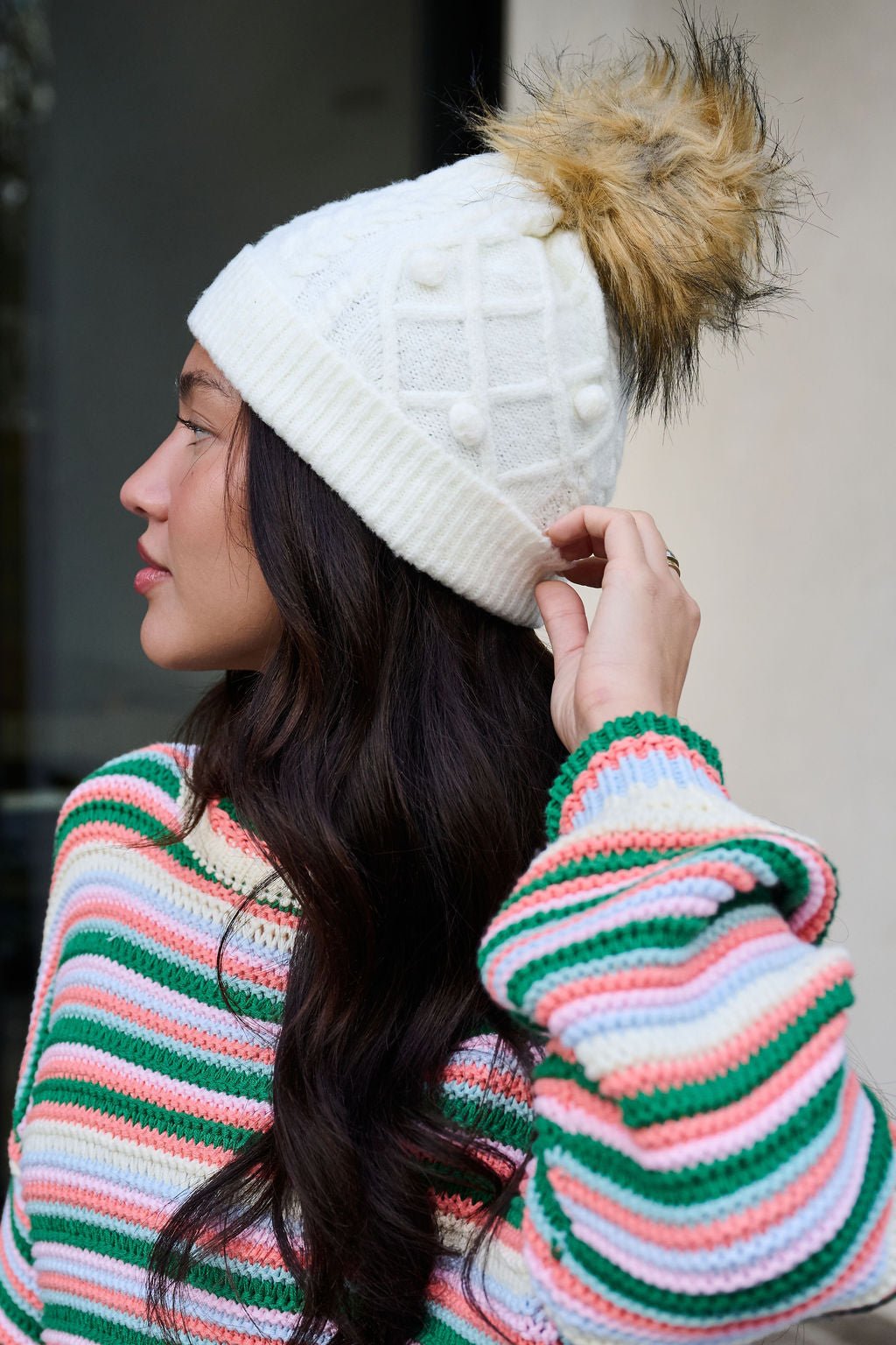 Woman in a colorful striped sweater wears the Fuzzy Ivory Cable Knit Pom Beanie, looking to the side.