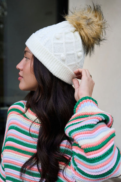 Woman in a colorful striped sweater wears the Fuzzy Ivory Cable Knit Pom Beanie, looking to the side.