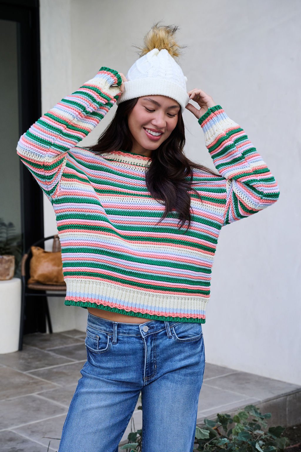 Woman smiling outdoors in a colorful striped sweater, blue jeans, and the Fuzzy Ivory Cable Knit Pom Beanie - FINAL SALE, hands on her hat.
