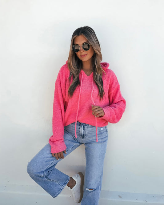 Woman wearing a Fuzzy Pink Hooded Sweater, light blue ripped jeans, white sneakers, and sunglasses stands by a white wall.