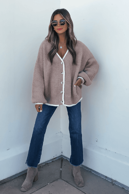 Woman in sunglasses, blue jeans, taupe boots, and a Fuzzy Soft Camel Button Down Cardigan stands by a white wall.