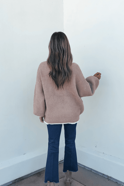 A woman with long wavy hair in a Fuzzy Soft Camel Button Down Cardigan and flared blue jeans stands facing a white wall.