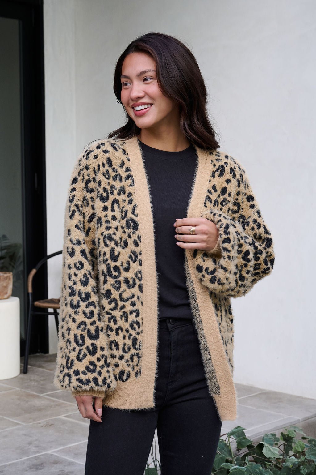 A woman smiles outdoors in a Fuzzy Tan Leopard Open Front Cardigan layered over a black shirt and pants.
