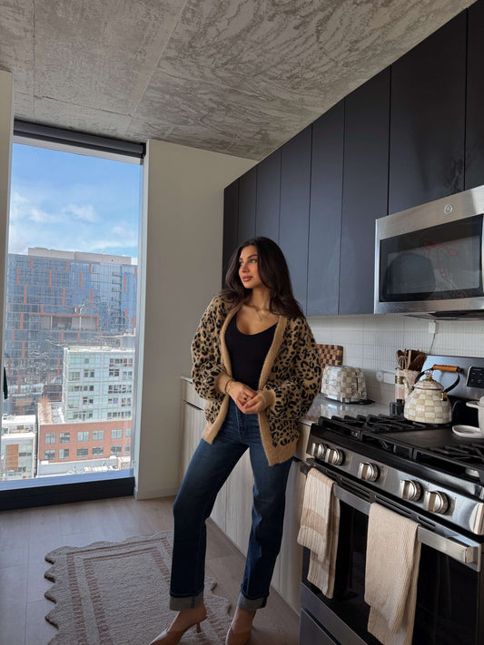 A woman in the Fuzzy Tan Leopard Open Front Cardigan stands by a stove in a modern kitchen with large windows and city views.