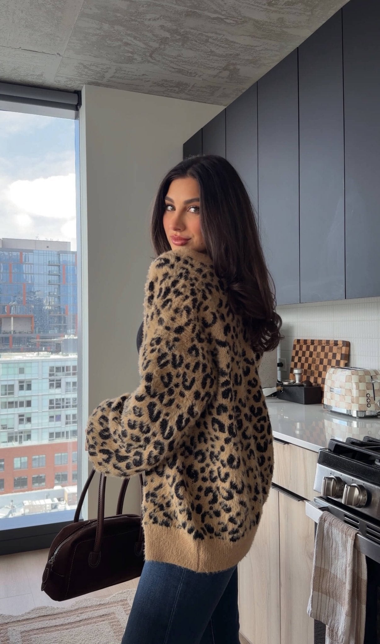 A woman wearing the Fuzzy Tan Leopard Open Front Cardigan stands in a modern kitchen, holding a brown bag.