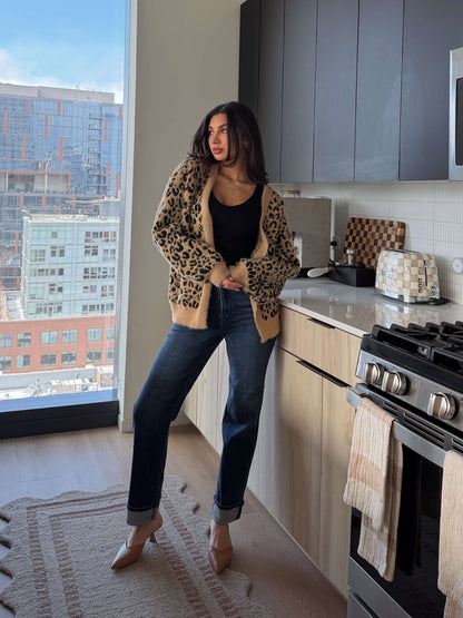 A woman in a Fuzzy Tan Leopard Open Front Cardigan stands in a modern kitchen with city buildings visible through the window.