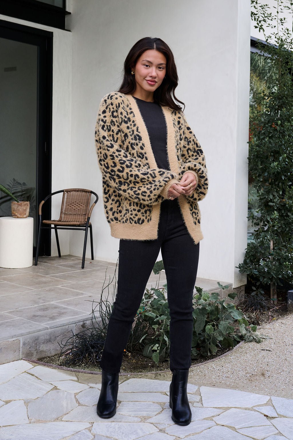 Woman outside in a Fuzzy Tan Leopard Open Front Cardigan, black top, jeans, and boots, near plants and a chair.