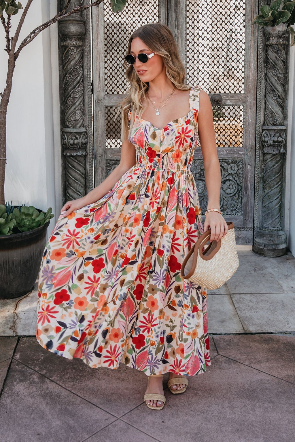 A woman in sunglasses wears the Garden Wayfarer Print Midi Dress with sandals and a woven bag, standing outdoors by ornate doors.