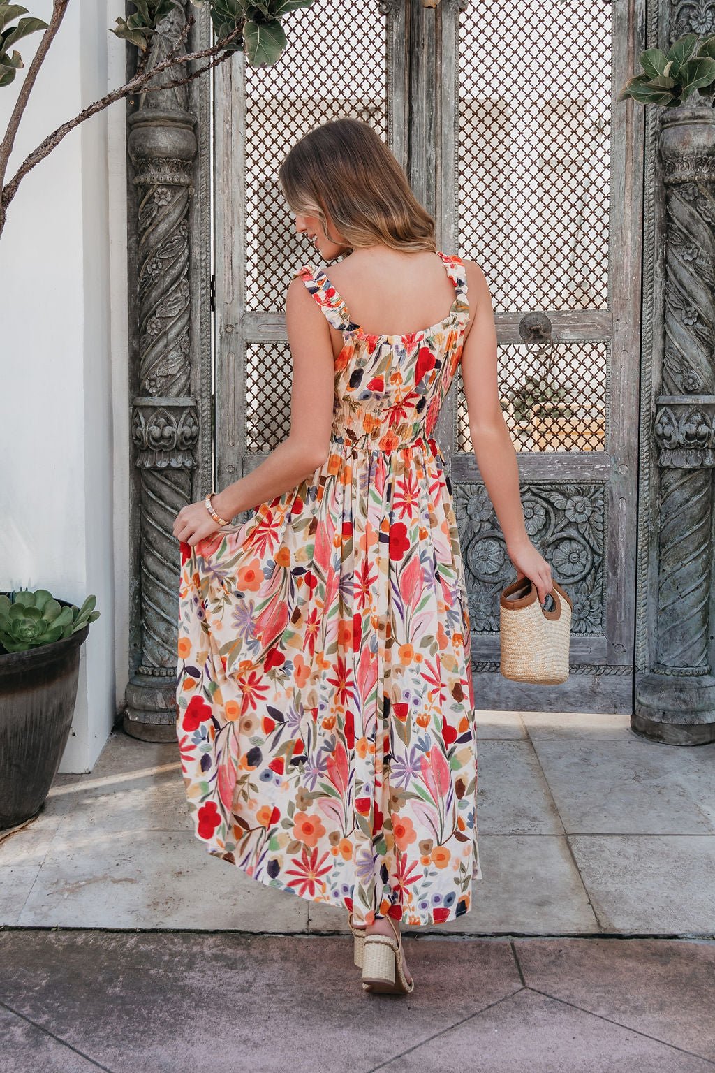 Woman in a Garden Wayfarer Print Midi Dress and sandals holds a woven handbag, standing on a patio before ornate wooden doors.