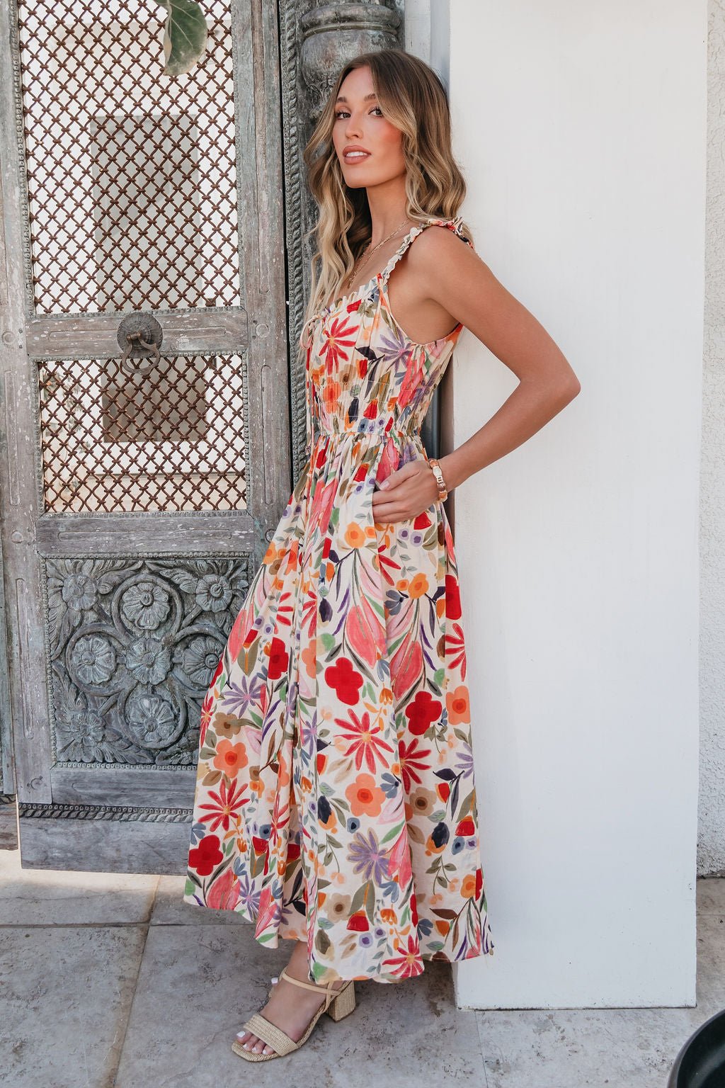Woman in a Garden Wayfarer Print Midi Dress and sandals stands by an ornate wooden door, looking to the side.