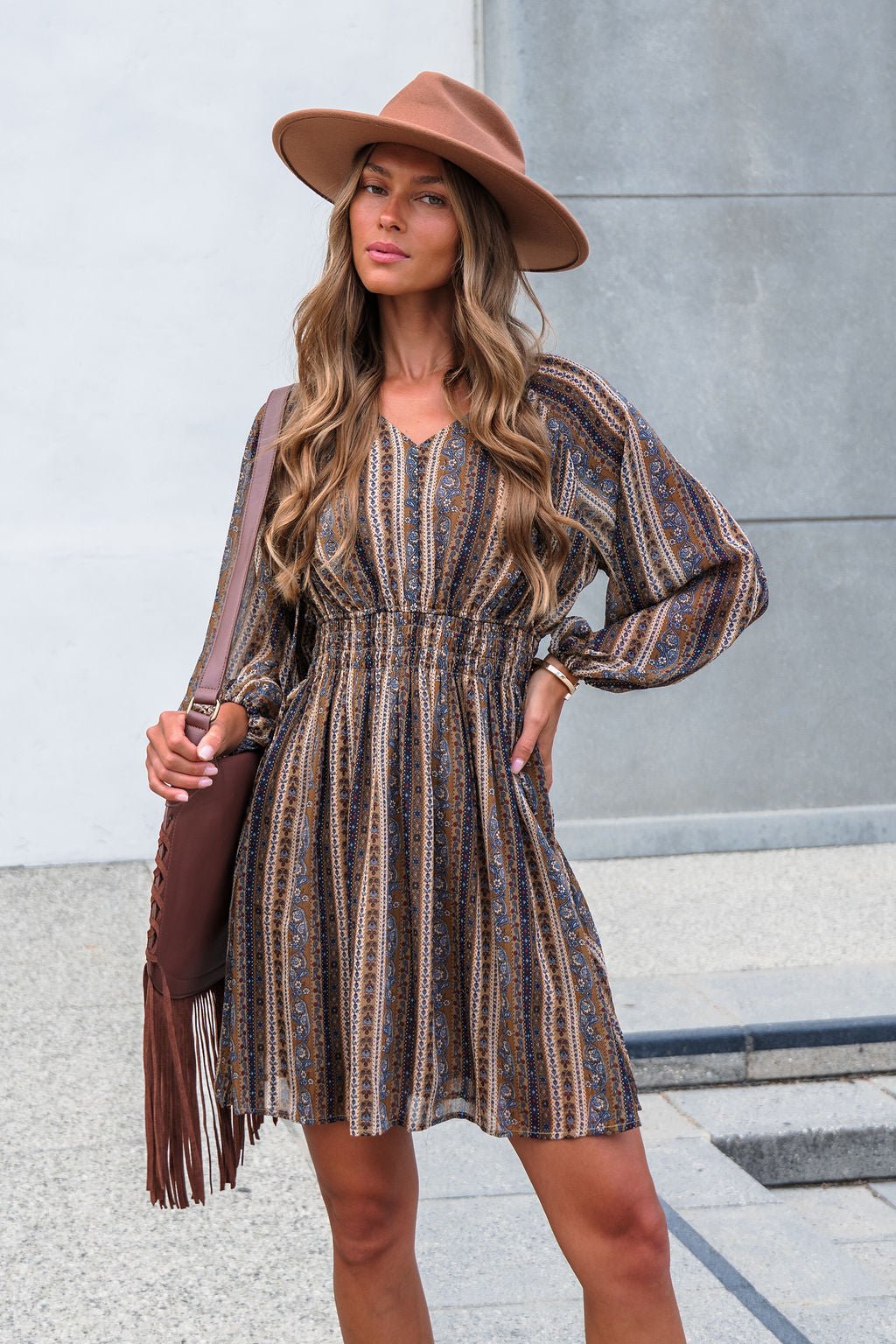 Woman in Genevieve Floral Striped Mini Dress—Mocha, with balloon sleeves, wide-brim hat, brown fringe bag, wavy hair, neutral expression.