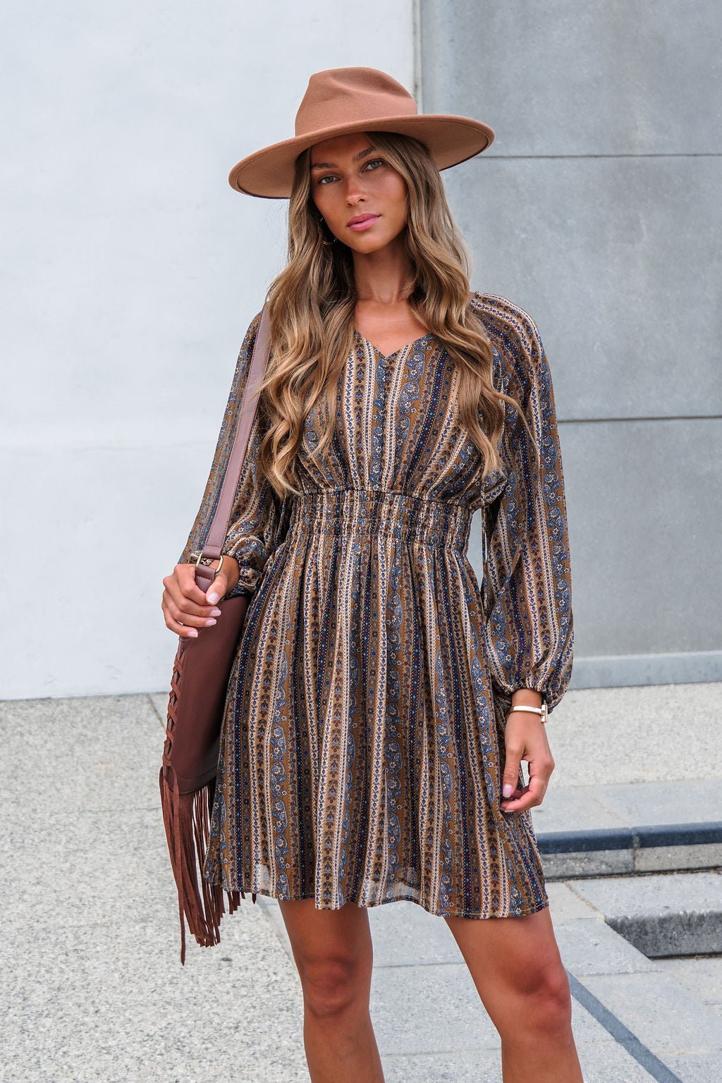 Woman in the Genevieve Floral Striped Mini Dress—Mocha, with balloon sleeves, poses outdoors with a wide-brim hat and fringe bag.