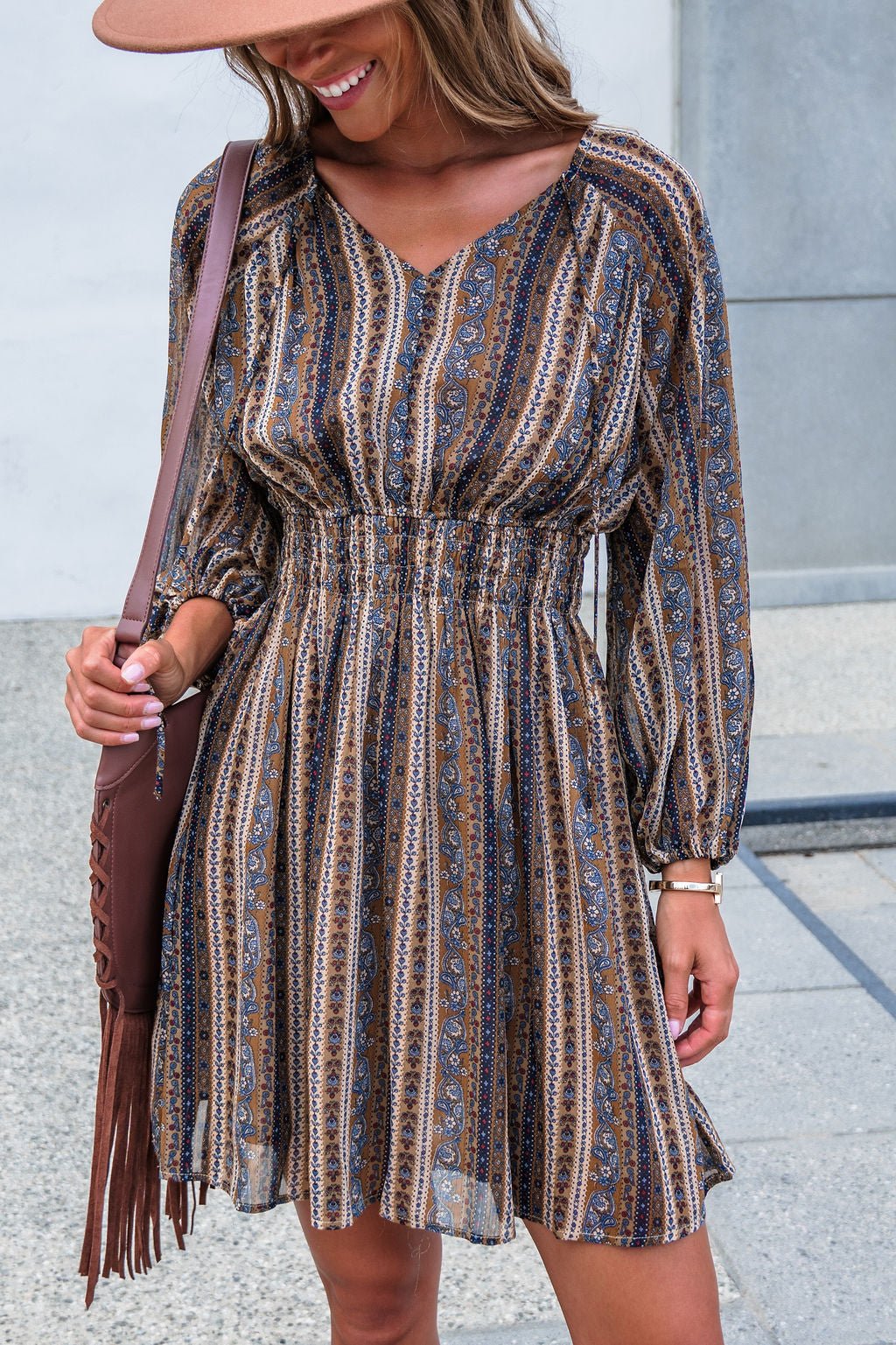 Smiling outdoors, woman in Genevieve Floral Striped Mini Dress — Mocha, with brown fringe bag and tan hat.