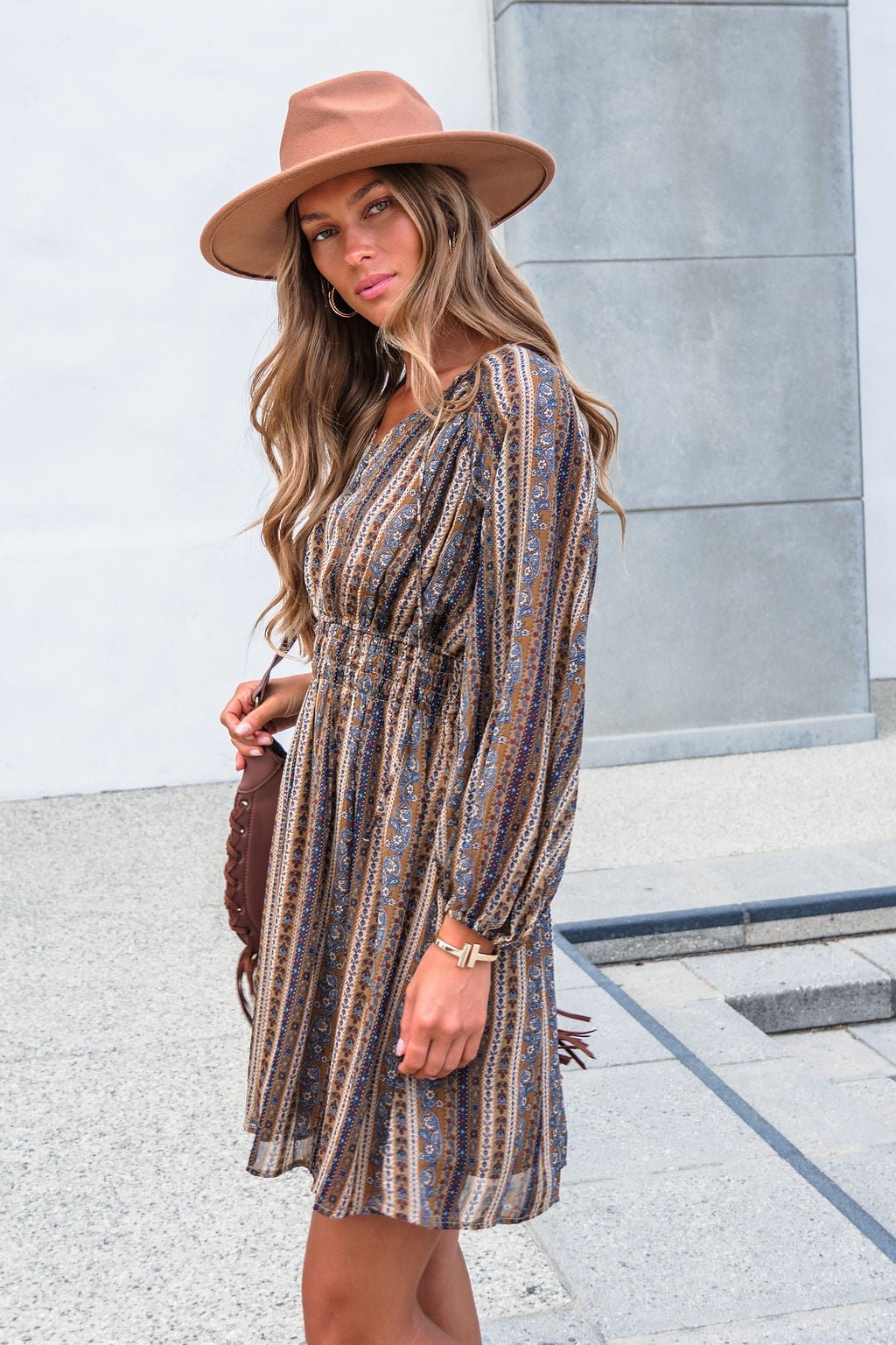 Woman wears the Genevieve Floral Striped Mini Dress—Mocha, with balloon sleeves and V-neck, standing outdoors holding a brown bag.