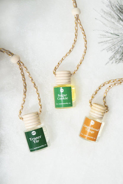 Three Gingerbread Village Car Diffusers rest on snow—festive glass bottles, perfect as car air fresheners or Christmas gifts.