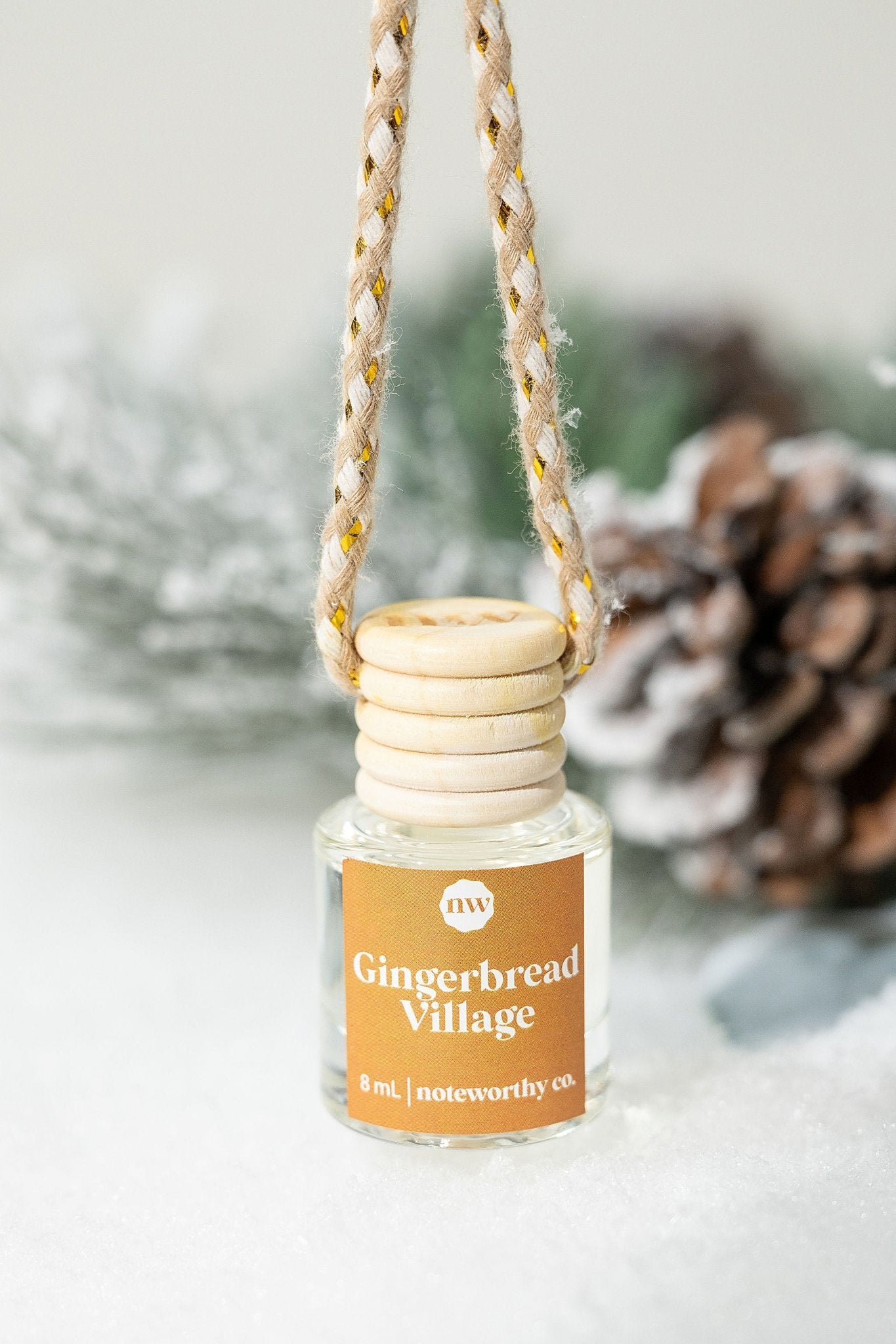 The Gingerbread Village Car Diffuser hangs on snow with pinecones and greenery—ideal as a Christmas gift.
