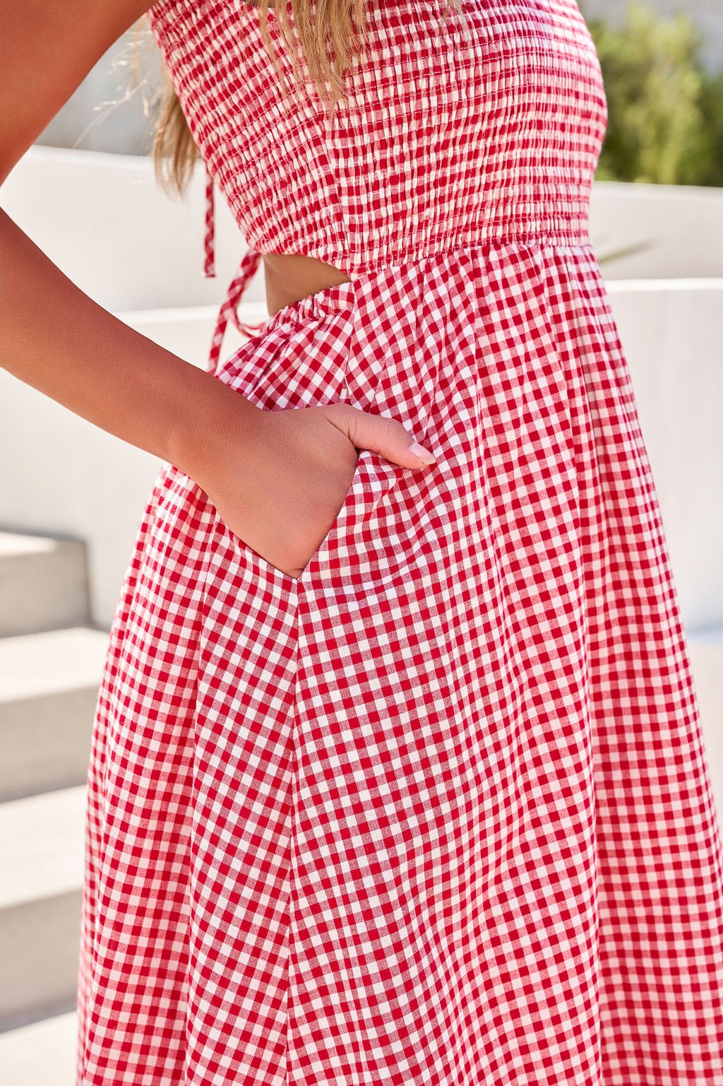 Woman in Gingham Open Back Midi Dress - Red, standing outdoors in sunlight with a hand in the pocket.