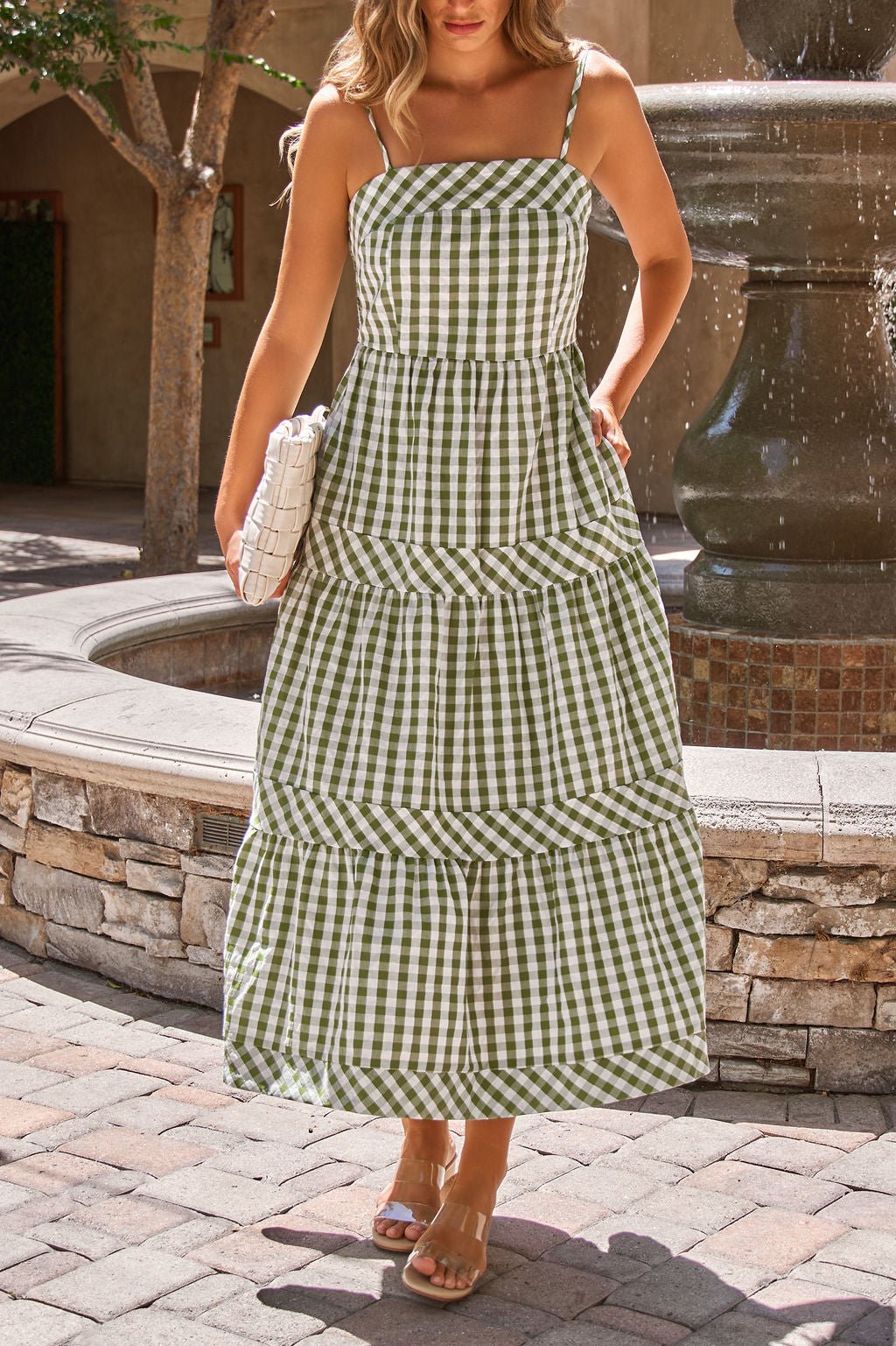 Woman in a Gingham Smocked Midi Dress - Olive, holding a white clutch, stands by a stone fountain.