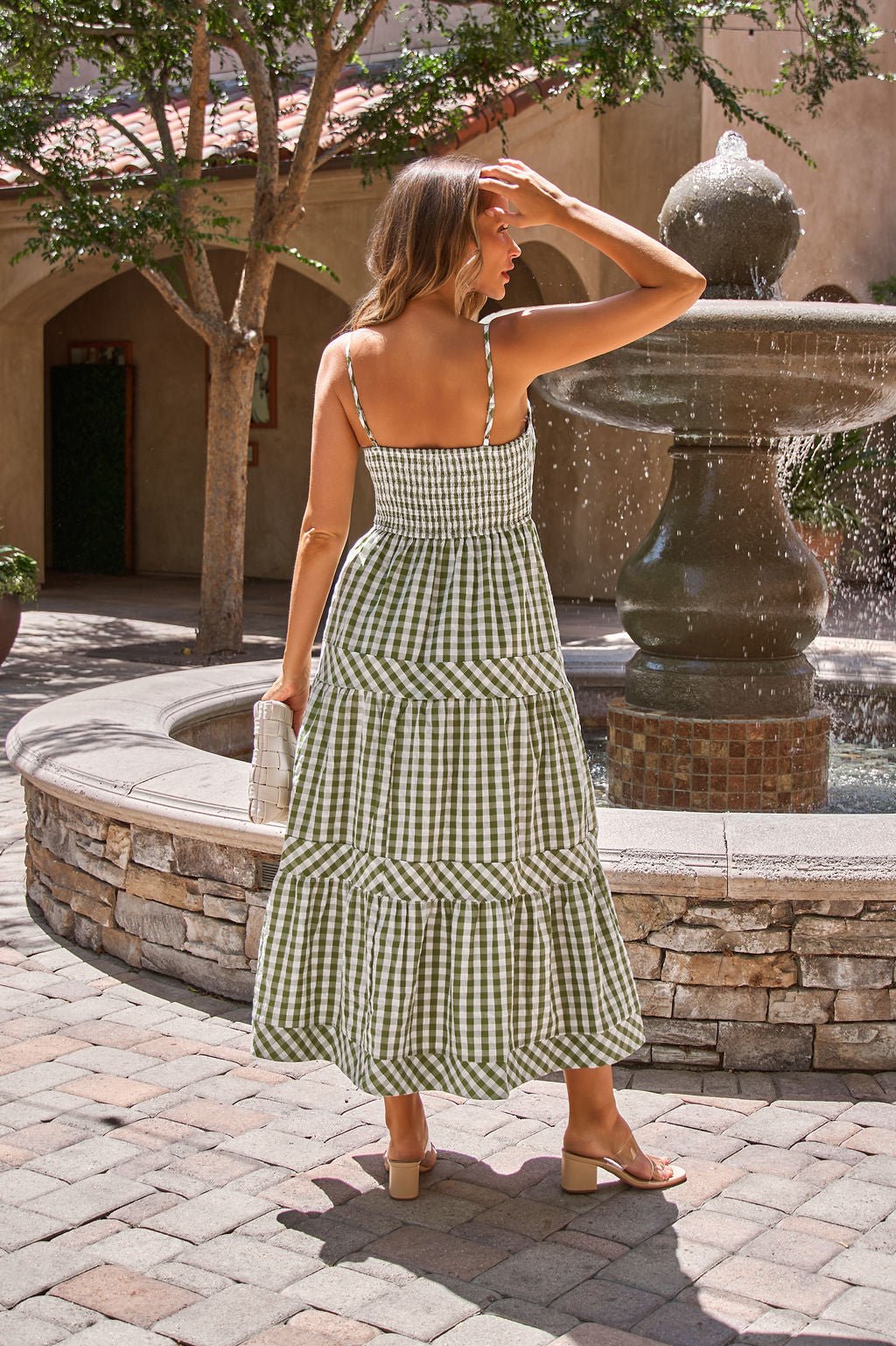 A woman in the Gingham Smocked Midi Dress - Olive stands by a stone fountain, clutch in hand, shielding her eyes from the sun.
