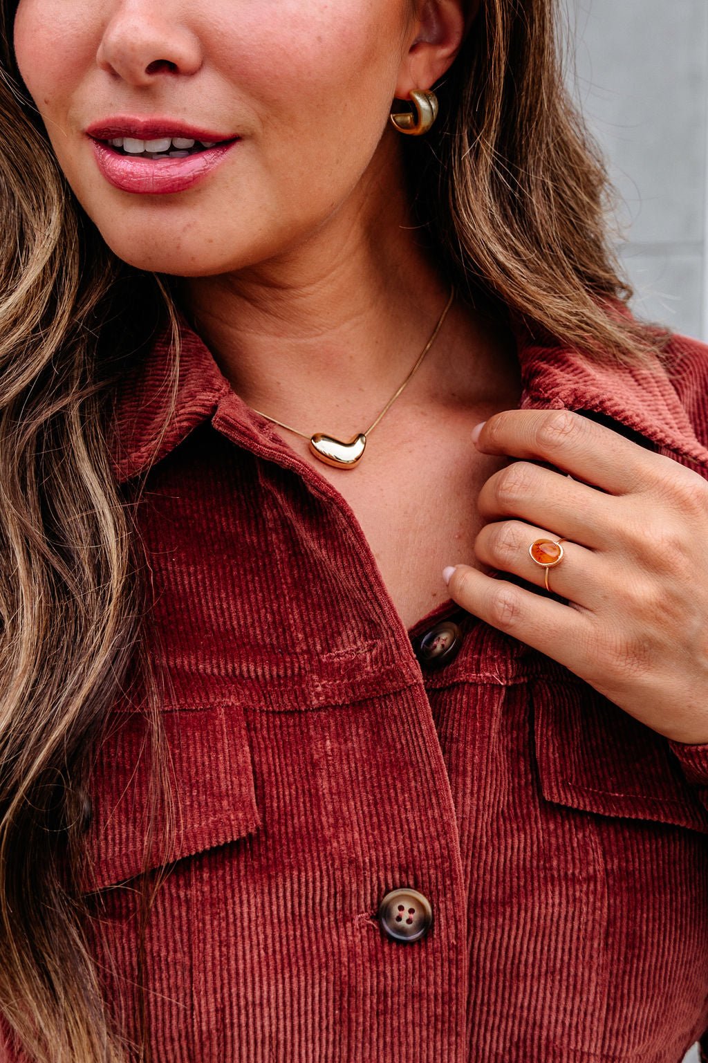 A woman in a rust corduroy shirt, gold hoops, and a Gold Abstract Heart Pendant Necklace, with wavy hair and an orange stone ring.