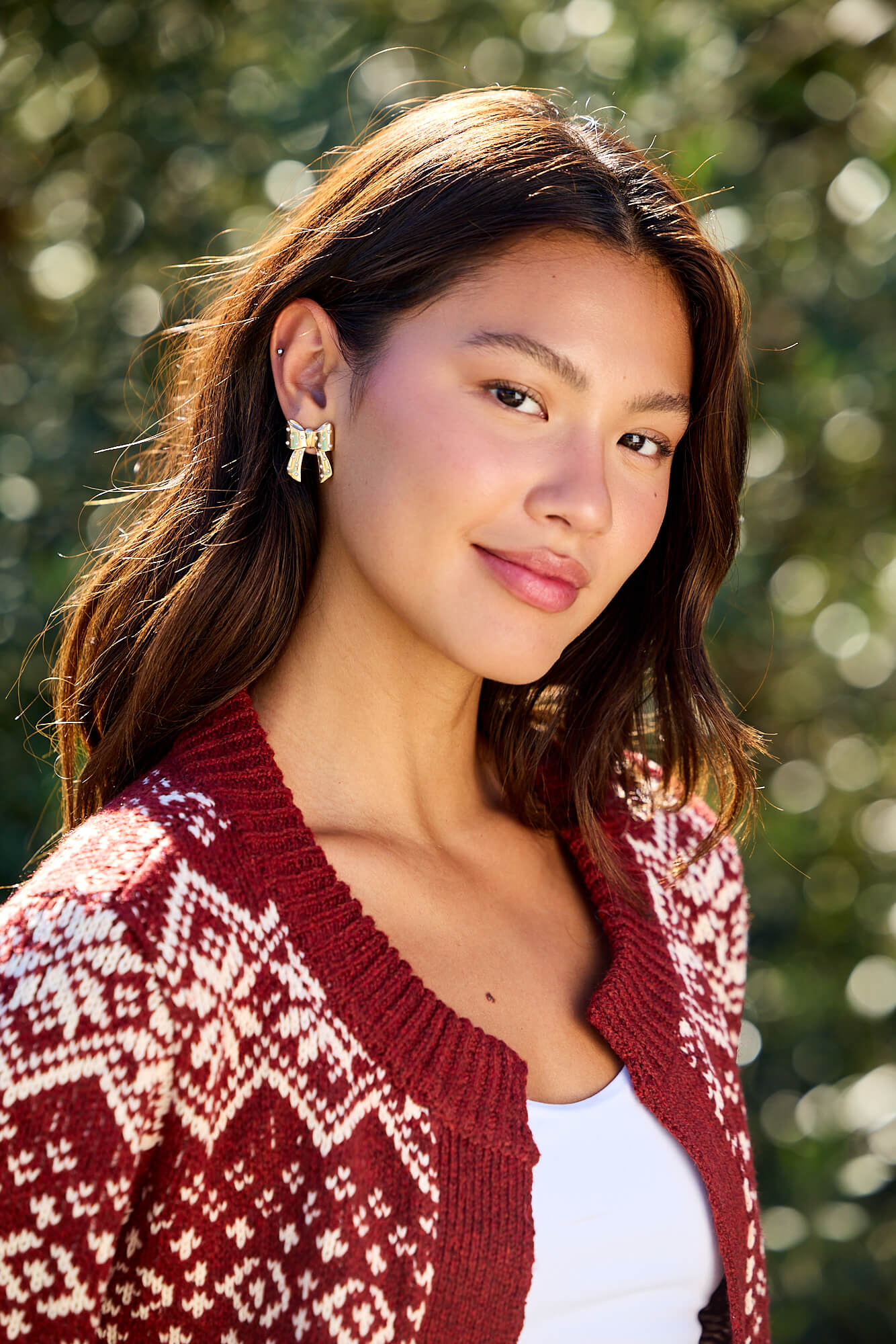 A woman with long brown hair smiles outdoors, wearing Gold and Pearl Bow Earrings and a red and white cardigan.