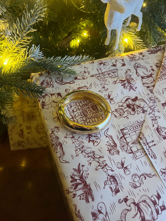 A Gold Beveled Hinge Bangle Bracelet sits on a gift with printed wrap beneath a Christmas tree and white horse ornament.