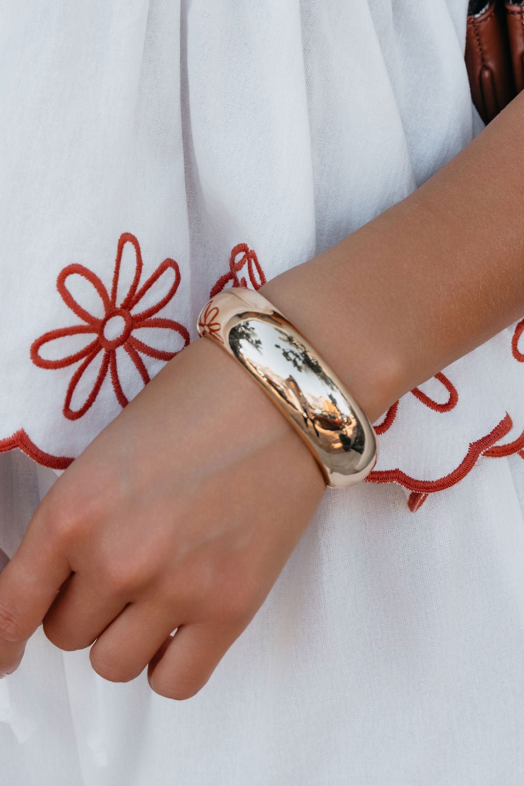 A hand showcases the Gold Beveled Hinge Bangle Bracelet on white fabric with red floral embroidery.