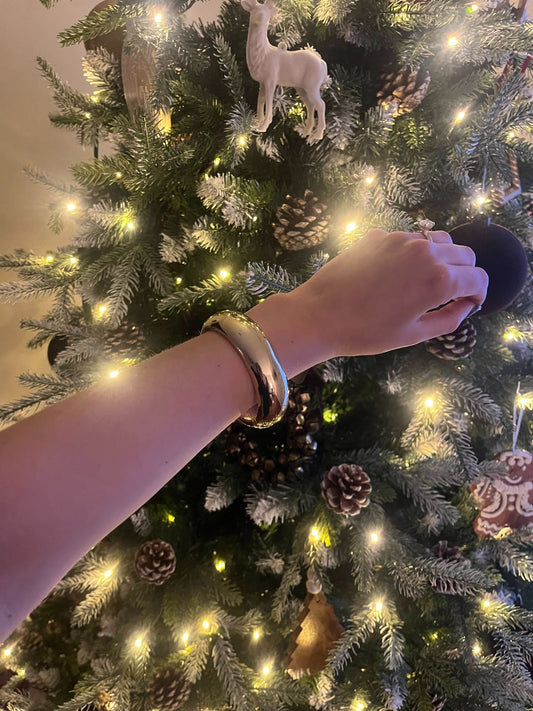 A hand models the Gold Beveled Hinge Bangle Bracelet in front of a decorated, lit Christmas tree with ornaments and pinecones.